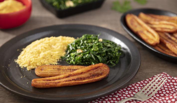 Imagem de um prato com um pedaço de banana frita, farofa e couve refogada. Ao lado, uma travessa com mais bananas fritas.