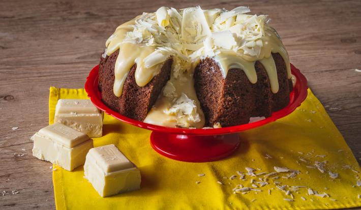 Fotografia em tons de amarelo, em uma bancada de madeira de cor marrom. Ao centro, um pano amarelo contendo o bolo vulcão. Ao lado, há 3 pedaços de chocolate branco e algumas raspas de chocolate espalhadas.
