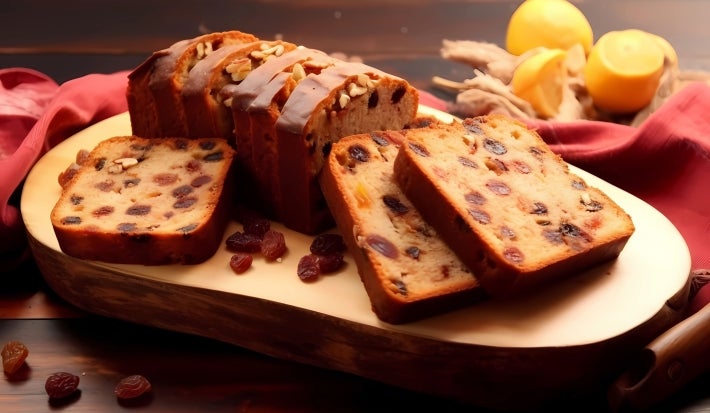 Fotografia de fatias de bolo de frutas cristalizadas dispostas em uma tábua de madeira, com textura rica e pedaços de frutas secas visíveis. O fundo apresenta um pano vermelho.