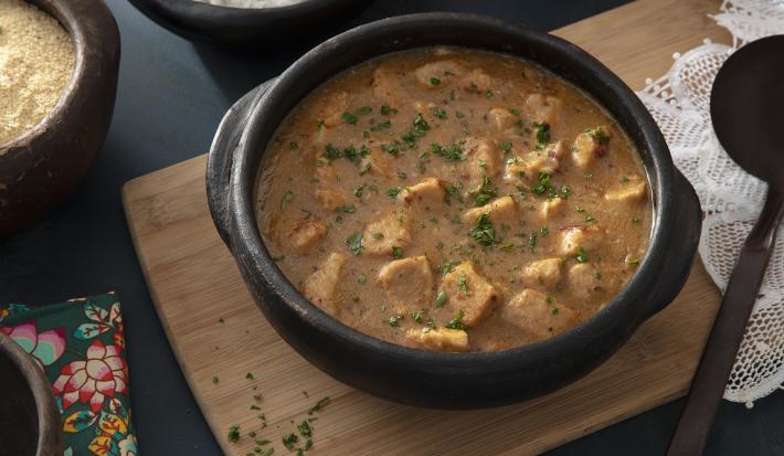 Foto da receita de Sarapatel. Observa-se uma panela de barro com a receita sobre uma tábua de madeira em uma superfície preta. Em cima, cumbucas de arroz e farofa.