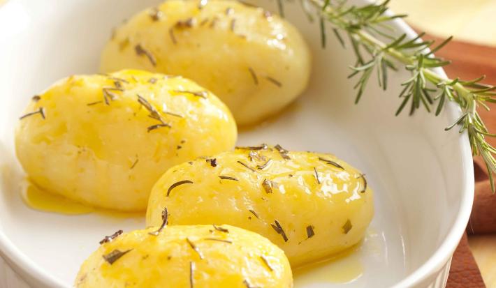 Fotografia em tons de branco e amarelo de uma bancada de madeira clara, com um pano marrom, um refratário branco oval com 4 batatas dentro, com azeite e alecrim e um ramo de alecrim.