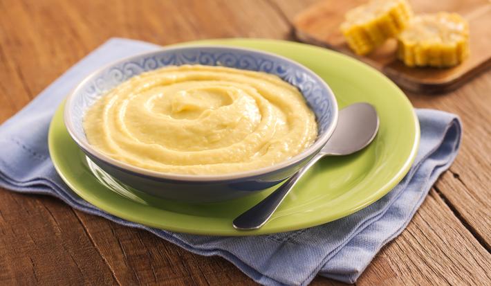 Fotografia em tons de verde e azul em uma bancada de madeira clara, com um paninho azul, um prato verde e outro azul fundo em cima com o creme de milho. Ao fundo, uma tábua de madeira com milhos cortados.