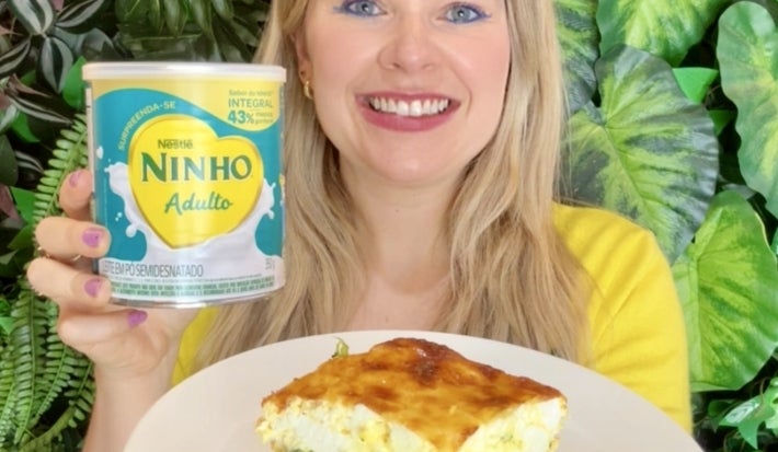 Fotografia em tons de verde com uma moça segurando um prato. No prato existe uma receita de torta de brócolis saudável feita com NINHO Adulto