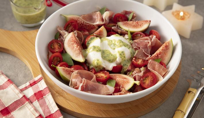Foto da receita de Burrata ao Pesto com Presunto Cru e Figos. Observa-se um recipiente refratário branco de porcelana redondo com a burrata dentro ao centro, e em volt os presuntos, figos e tomates. Observa-se molho pesto regado por cima.