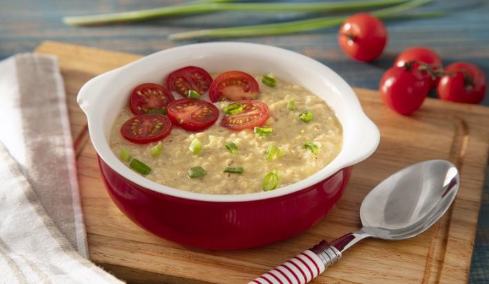 Foto em tons de vermelho da receita de aveia salgada servida em uma panelinha de cerâmica vermelha sobre uma tábua de madeira com uma colher prateada ao lado. Ao fundo temos alguns tomates-cereja