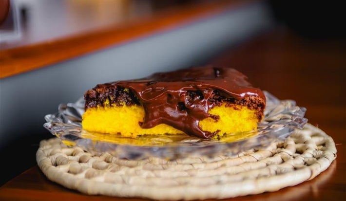 Fotografia de uma fatia de bolo de cenoura com brownie por cima e uma calda de chocolate brilhante para finalizar. A fatia está em um prato de vidro pequeno, apoiado em um apoio raso de bambu de tom claro.