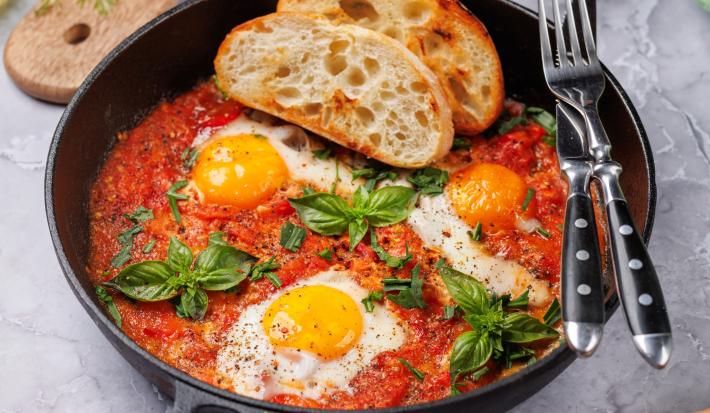 Fotografia em tons claros de uma receita de Shakshuka com pães por cima, dentro de uma frigideira de ferro preta sobre uma mesa cinza. Tem um par de talheres apoiados na frigideira, que está sobre uma mesa cinza.
