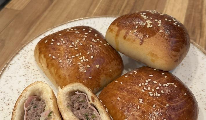 Fotografia de três hambúrgueres de forno recheados com presunto e queijo sobre um prato branco apoiado em uma tábua de madeira. Ao lado dos hambúrgueres tem mais um, mas que está aberto ao meio.