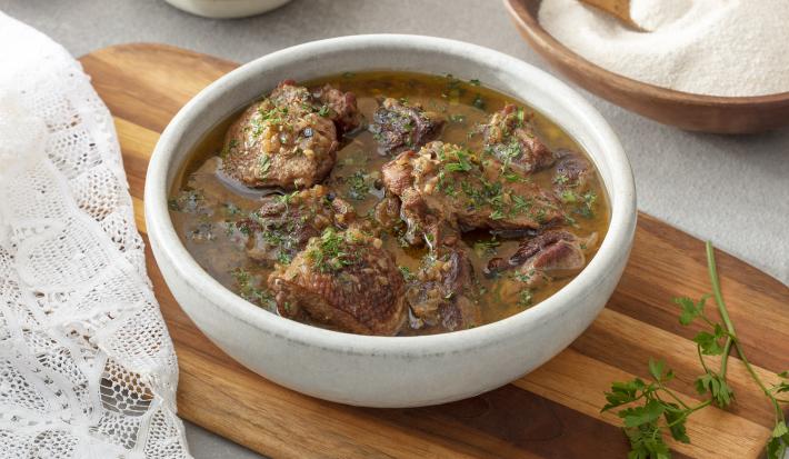 Foto em tons de marrom da receita de pato no tucupi servida em um bowl de cerâmica branco sobre uma tábua de madeira com cheiro verde ao lado. Ao fundo há um recipiente de madeira com farinha de mandioca e ao lado um paninho branco