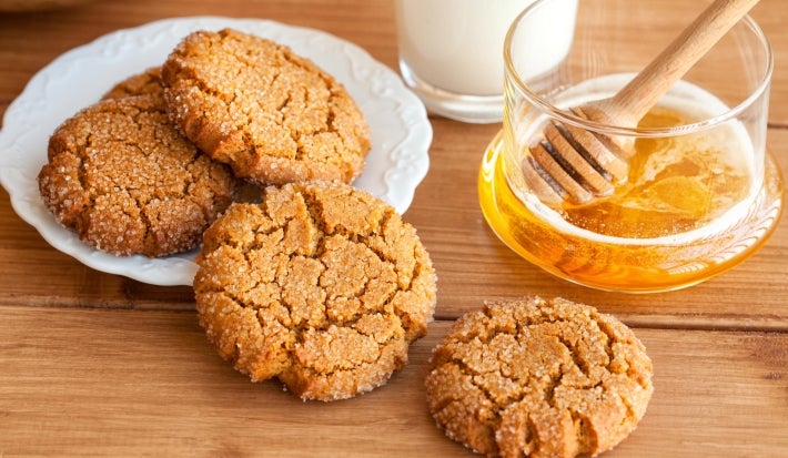 Em cima de uma mesa, prato branco com biscoitos crocantes, decorados com granulado de açúcar cristal. Ao lado do prato, pote com mel.