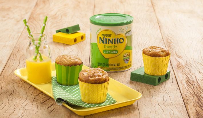 Fotografia em tons de amarelo e verde em uma bancada de madeira clara, um recipiente amarelo com dois cupcakes de maçã e aveia em forminhas amarela e verde. Ao lado, brinquedinhos infantis, uma lata de Ninho Fases e mais um cupacke.