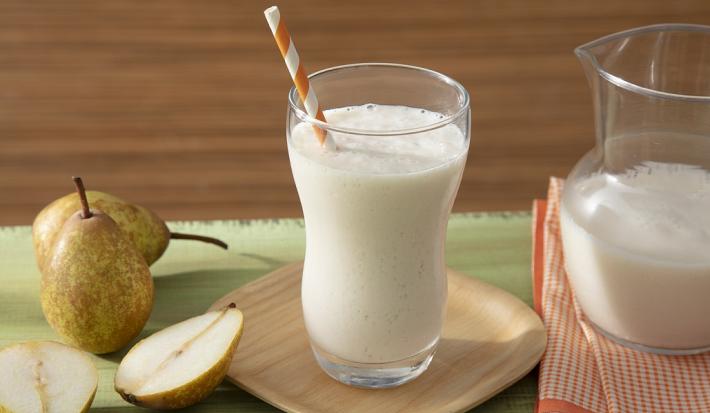 Fotografia em tons de branco em uma bancada de madeira de cor verde, ao centro, um pires de madeira contendo um copo com o smoothie. Ao lado, há algumas peras e do outro lado, um pano laranja quadriculado com uma jarra em cima.