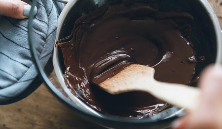 Fotografia vista de cima de uma mão feminina segurando uma colher de madeira e mexendo uma cobertura em banho-maria, feita de chocolate meio amargo e café. Os recipientes do banho-maria estão sobre uma bancada de madeira.
