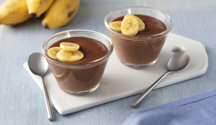 Fotografia em tons de azul e marrom de uma bancada com um paninho azul, duas tacinhas transparentes com o creme de chocolate e rodelas de banana decorando, ao lado duas colheres pratas e ao fundo 3 bananas.