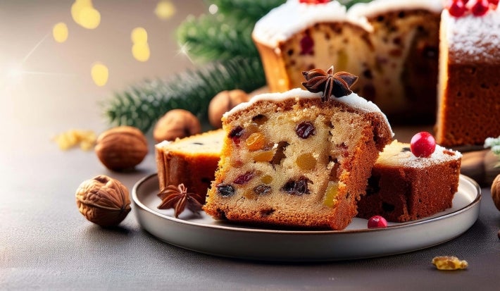 Fotografia de um bolo de frutas cristalizadas, cortado, com textura densa e recheado de frutas secas coloridas. Polvilhado com açúcar de confeiteiro, é decorado com anis estrelado e uma baga vermelha.