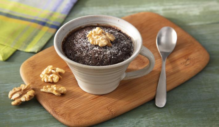 Fotografia em tons de verde em uma bancada de madeira verde, uma tábua de madeira, uma caneca bege com o brownie de chocolate com nozes dentro dela. Ao lado, nozes caídas e um paninho verde.