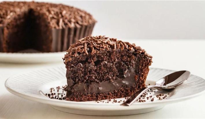 Fotografia em tons claros de uma fatia grande de bolo de chocolate molhadinho com brigadeiro e granulado por cima ao lado de uma colher, sobre um prato raso branco.