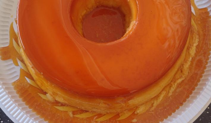 Fotografia de um pudim inteiro com caramelo dentro de um recipiente fundo branco de plástico.