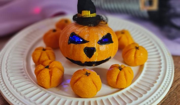Fotografia em tons de laranja, em uma bancada de madeira, um prato branco ao centro com os beijinhos em formato de abóbora. No centro, uma abóbora pequena com desenho de Halloween e um chapeuzinho de bruxa ao fundo da foto.