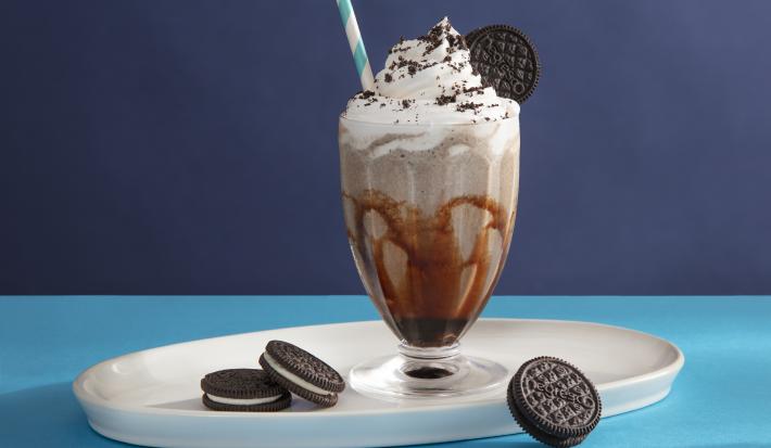 Foto da receita de Milkshake de negresco. Observa-se um copo de milkshake de vidro transparente decorado com calda e com a bebida dentro e topping de chantilly e farofa de biscoito, com um canudo azul e branco espetado