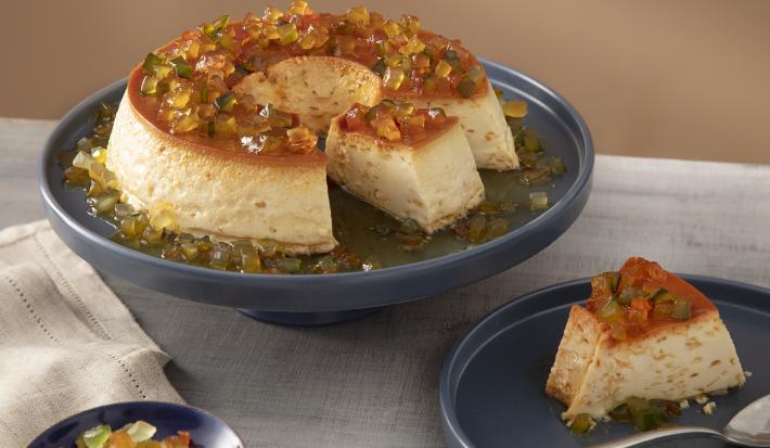 Foto da Receita de Pudim com Frutas Cristalizadas. Observa-se um recipiente de servir azul marinho com o pudim e, ao lado direito, um prato individual com uma fatia e uma colher.