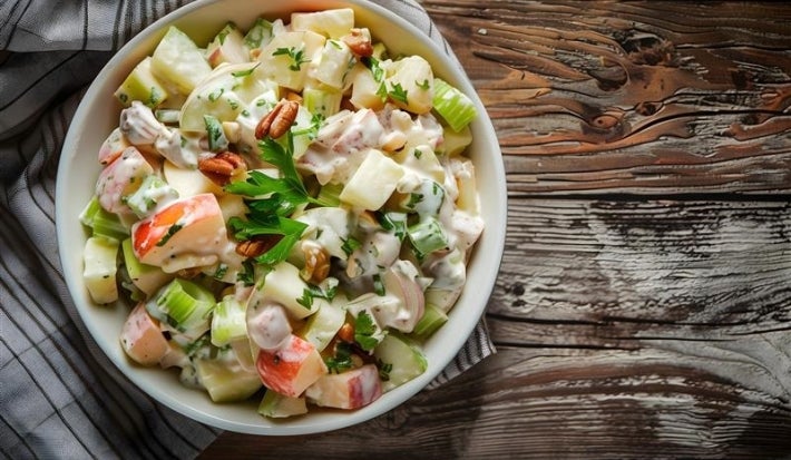 Fotografia de uma salada fresca em uma tigela branca sobre uma mesa de madeira rústica. A salada contém fatias de maçã, batatas cozidas, nozes, pedaços de maçã, um molho cremoso e salsa, apresentando um visual apetitoso.