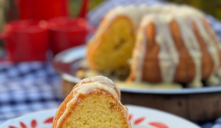 Fotografia de um pedaço de bolo de laranja com brigadeiro de laranja em um prato raso branco grande. Ao fundo tem o restante do bolo com a calda.