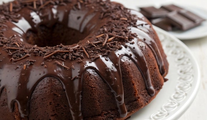 Fotografia de um bolo de chocolate em forma de bundt, coberto com um brilho de chocolate que escorre pelas laterais. Decorado com raspas de chocolate, está servido em um prato branco, com um fundo desfocado de pedaços de chocolate escuro.