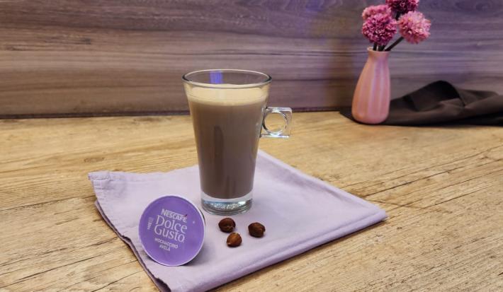 Fotografia em tons de lilás em uma bancada de madeira, com um paninho lilás, uma xícara de vidro alta com o Mochaccino Avelã dentro dela, ao lado, uma cápsula de Dolce Gusto e ao fundo um vaso de flor e um paninho marrom.