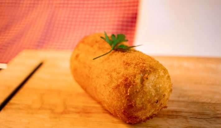 Fotografia de um croquete frito de carne moída sobre uma tábua de madeira. Tem um pequeno ramo de salsinha decorando o croquete.