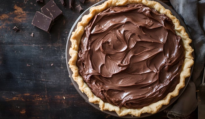 Fotografia vista de cima em tons escuros de uma torta com cobertura de chocolate sobre um praot grande e raso, apoiado em uma mesa de madeira escura.
