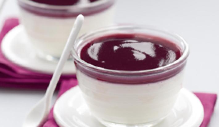 Fotografia em tons de roxo escuro em uma bancada de madeira cinza, dois guardanapos de pano roxos, dois pratinhos de café brancos e dois copinhos de vidro com a sobremesa pannacotta com calda de vinho tinto dentro.