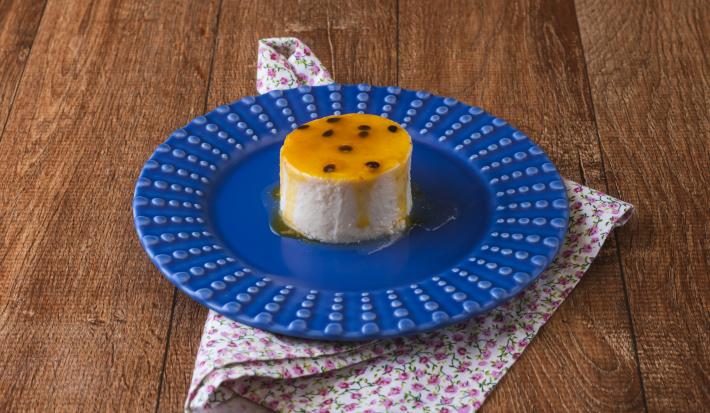 imagem da receita de pudim de iogurte da rafaela masaia, servido em um prato azul com uma calda de frutas amarelas. O prato está sobre um pano florido em tom rosa em uma bancada de madeira