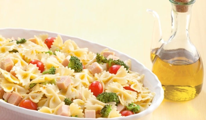 Travessa branca com salada de macarrão gravatinha com peito de peru, brócolis e tomate cereja com um vidro de azeite ao lado.