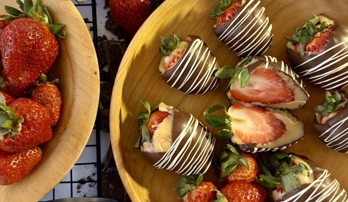 Fotograifa mostra bombons de morango sobre um prato de madeira. É possível ver um dos bombons abertos, mostrando a camada de chocolate, morango e brigadeiro.