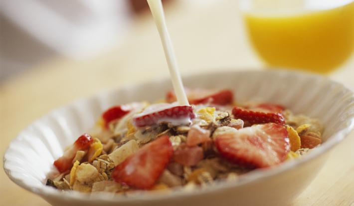 Fotografia de um recipiente branco de vidro fundo um creme com frutas e cereal Nesfit. O recipiente está sobre uma mesa de madeira, e ao fundo tem um copo com suco de laranja.