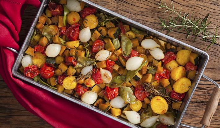 Fotografia de um recipiente de ferro com alças de madeira, dentro estão todos os legumes ficados em cubos assados, e ao lado está um guardanapo de tecido vermelho.