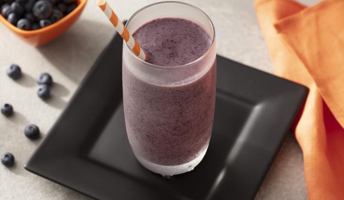 Fotografia em tons de preto, laranja e branco de uma bancada de madeira, sobre ela um prato quadrado preto com um copo de smoothie. Ao lado um paninho laranja e um recipiente laranja com mirtilo.