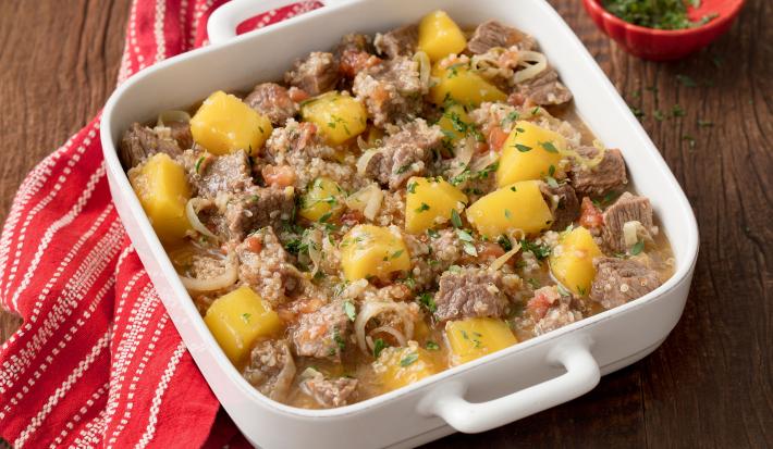 Fotografia em tons de marrom e branco uma bancada vista de cima, ao centro um recipiente quadrado com alças cor branco, dentro dele cubos de carne e pedaços de Mandioquinha e Quinoa. Ao lado um pano listrado na cor vermelho e branco.