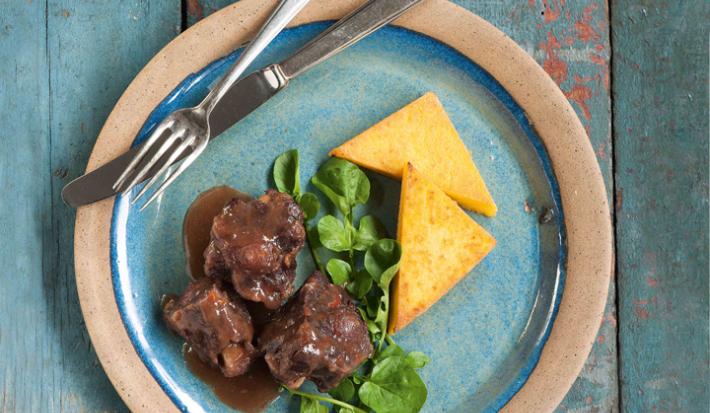Fotografia em tons de azul em uma bancada de madeira azul com um prato redondo raso rosa claro com azul e a rabada, a polenta e salada de agrião em cima dele.