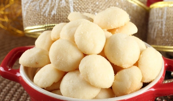 Em cima de uma mesa, recipiente redondo com biscoitos amanteigados redondos e dourados.