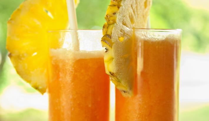 Fotografia de dois copos de vidro grandes com suco de laranja, abacaxi e mel. Em cada copo, tem metade de uma fatia de abacaxi e tem também um canudo. Ao fundo tem uma paisagem com de natureza com árvores.