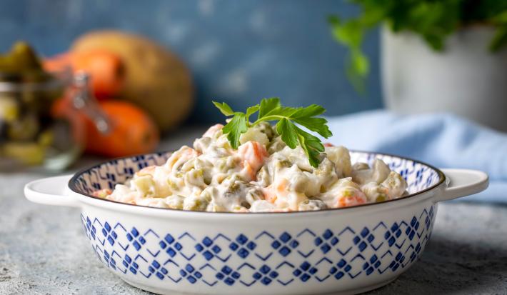 Fotografia de uma salada de maionese que está em um recipiente fundo com desenhos azuis. Tem um ramo de salsinha apoiado na salada. O recipiente está apoiado em uma bancada de textura parecida com gesso, ao fundo tem frutas.