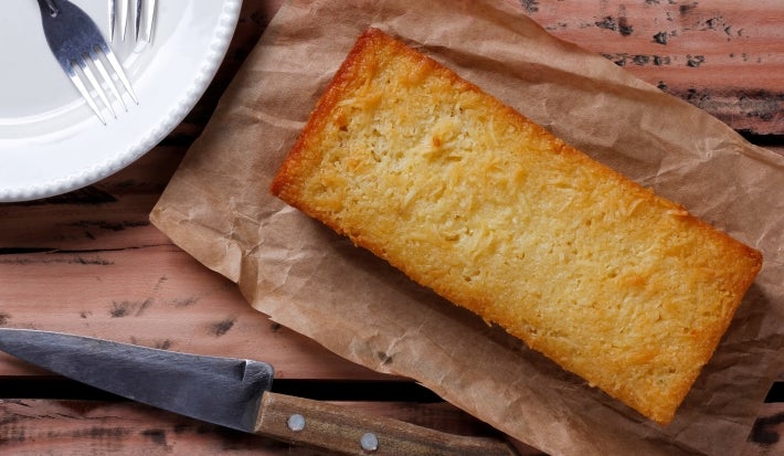 Um bolo de mandioca dourado e úmido, em formato retangular, está colocado sobre um papel manteiga. Ao lado, há uma faca rústica de madeira e um prato branco com garfos. O bolo tem aparência caseira e textura levemente crocante nas bordas.