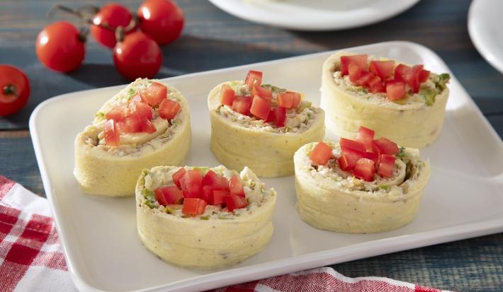 Foto em tons claros da receita de rolinho de ovos servida em 5 porções sobre uma base de porcelana branca. Ao fundo há um ramo de tomates-cereja e ao lado um paninho quadriculado vermelho e branco