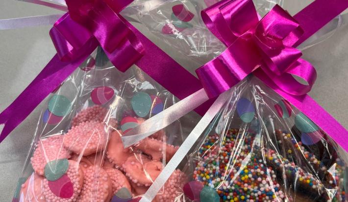 Imagem de dois saquinhos plásticos com laço grande cor de rosa sobre cada um deles, apoiados em um fundo claro. Dentro dos saquinhos, pequenas porções de chocolate cor de rosa e marrom com confeitos coloridos.