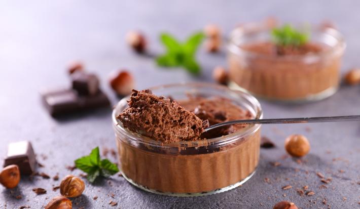 Imagem de um potinho redondo de vidro com mousse de chocolate e uma colher mostrando a textura cremosa da receita. Ao fundo, pedaços de chocolate e folhas de hortelã decorando.