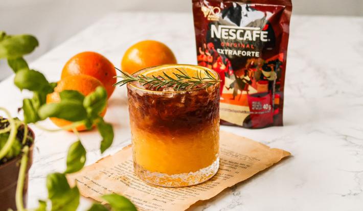 Fotografia em tons de laranja em uma bancada de mármore com um copo de vidro ao centro e o suco de laranja com café dentro dele, decorado com um ramo de alecrim. Ao fundo, laranja e embalagem de café.