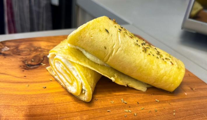Fotografia em tons de amarelo com uma tábua de madeira ao centro. Em cima da tábua existe uma crepioca recheada com creme de ricota e Ninho Adulto