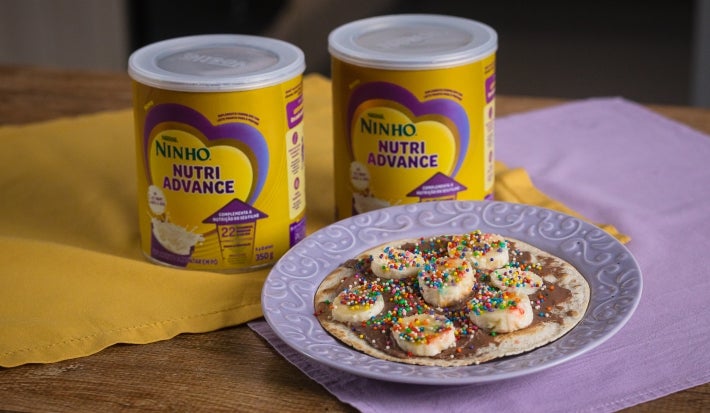Fotografia de dois pano, roxo e amarelo, em uma mesa de madeira. Sobre esses panos tem duas latas de NINHO Nutri Advance ao lado de um prato roxo claro com uma mini pizza com chocolate e banana.